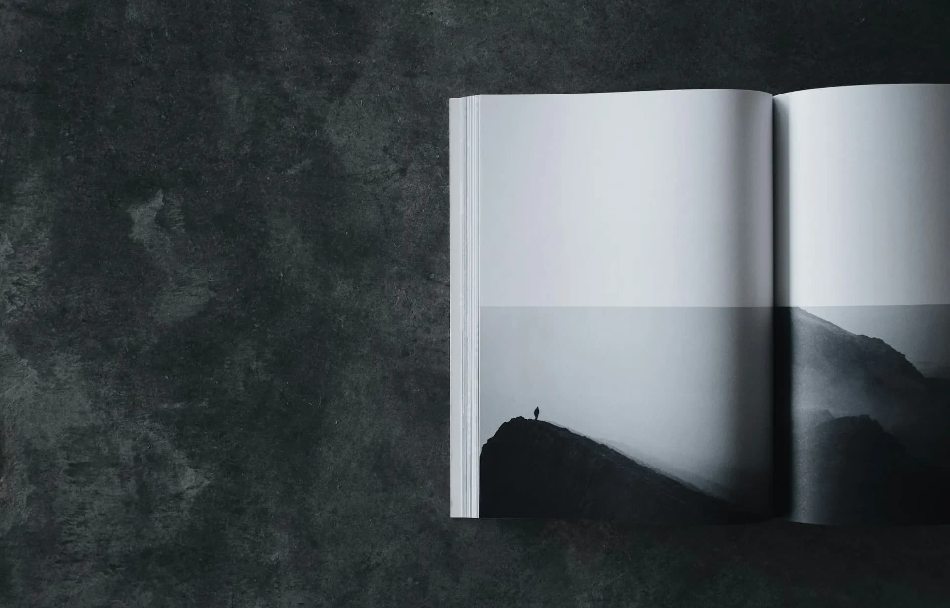 Open book displaying a black and white photo of a lone figure on a mountain ridge, placed on a dark textured surface.
