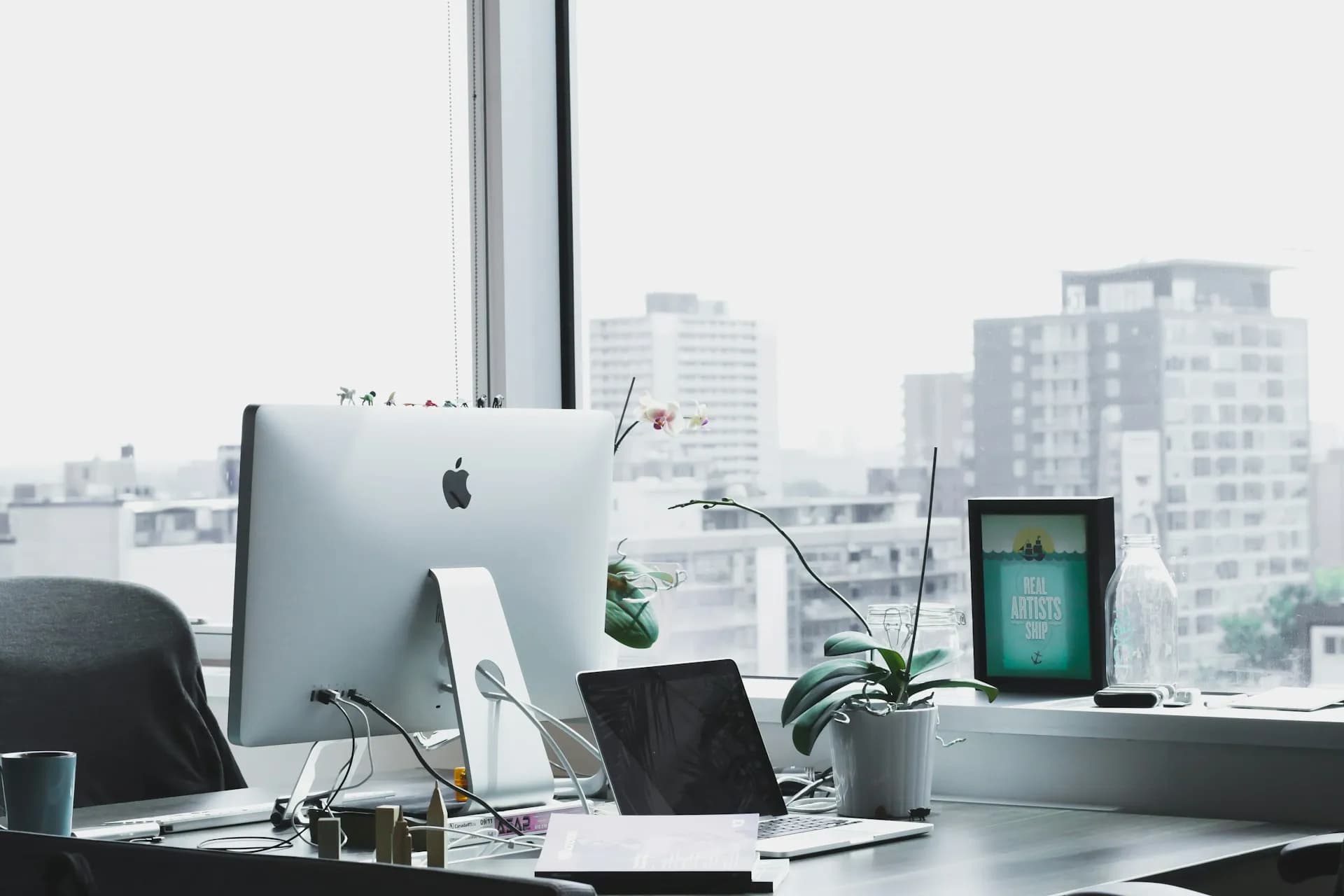 Modern office desk setup with an iMac, laptop, potted plants, and framed artwork, positioned near a window overlooking a cityscape.