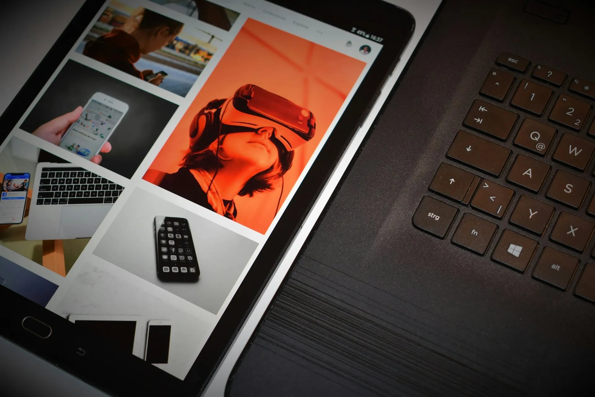 A tablet displaying a photo gallery, including an image of someone in a VR headset, lies next to a black laptop.