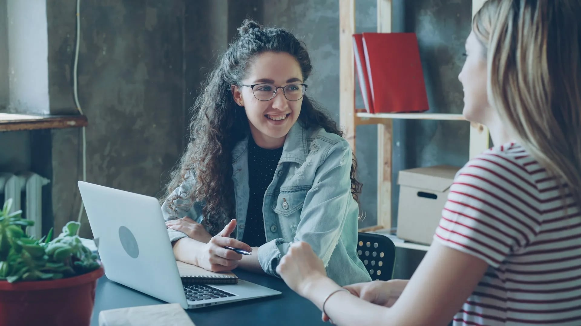 Two women having a friendly discussion at a desk with a laptop in a modern office setting.
