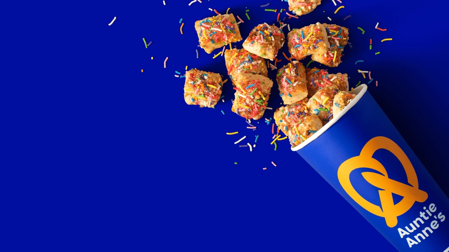 A cup of Auntie Anne's pretzel nuggets topped with colorful sprinkles, spilling onto a bright blue background.