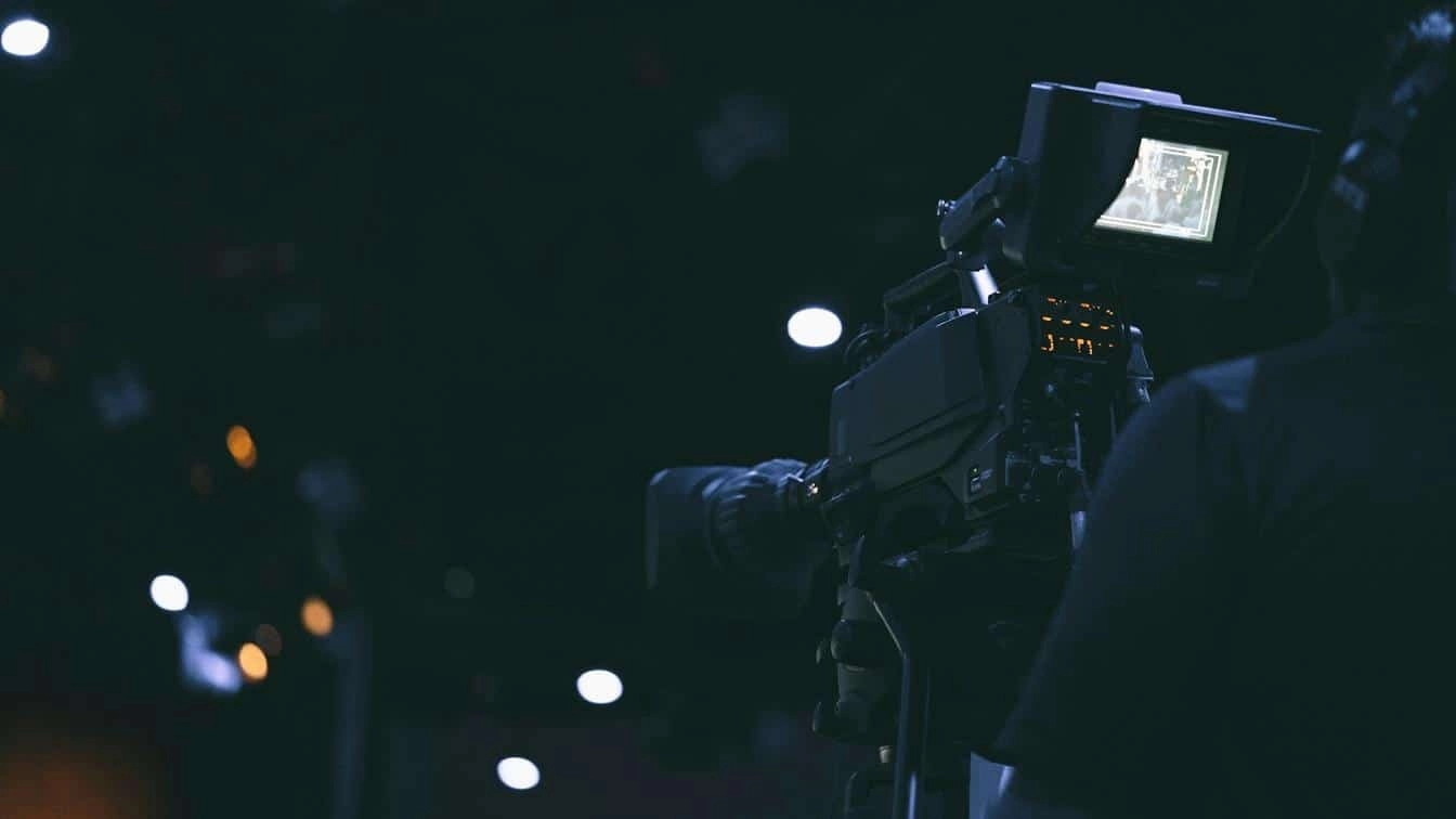 Operator behind a professional broadcast camera capturing media content in a dark studio setting.