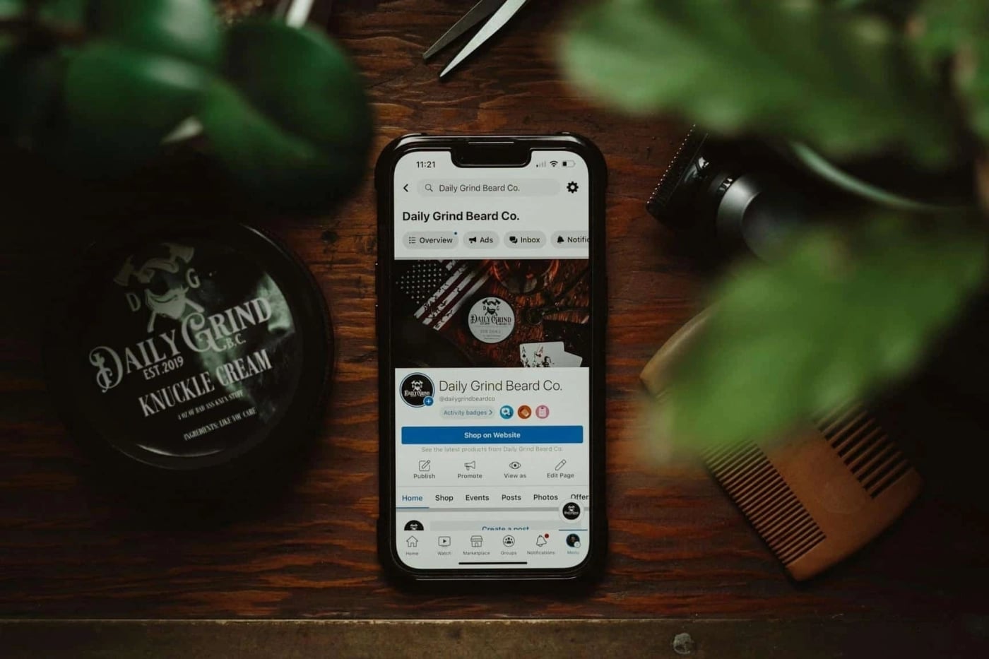 Smartphone displaying Daily Grind Beard Co. Facebook page on a wooden table with beard care products and accessories.