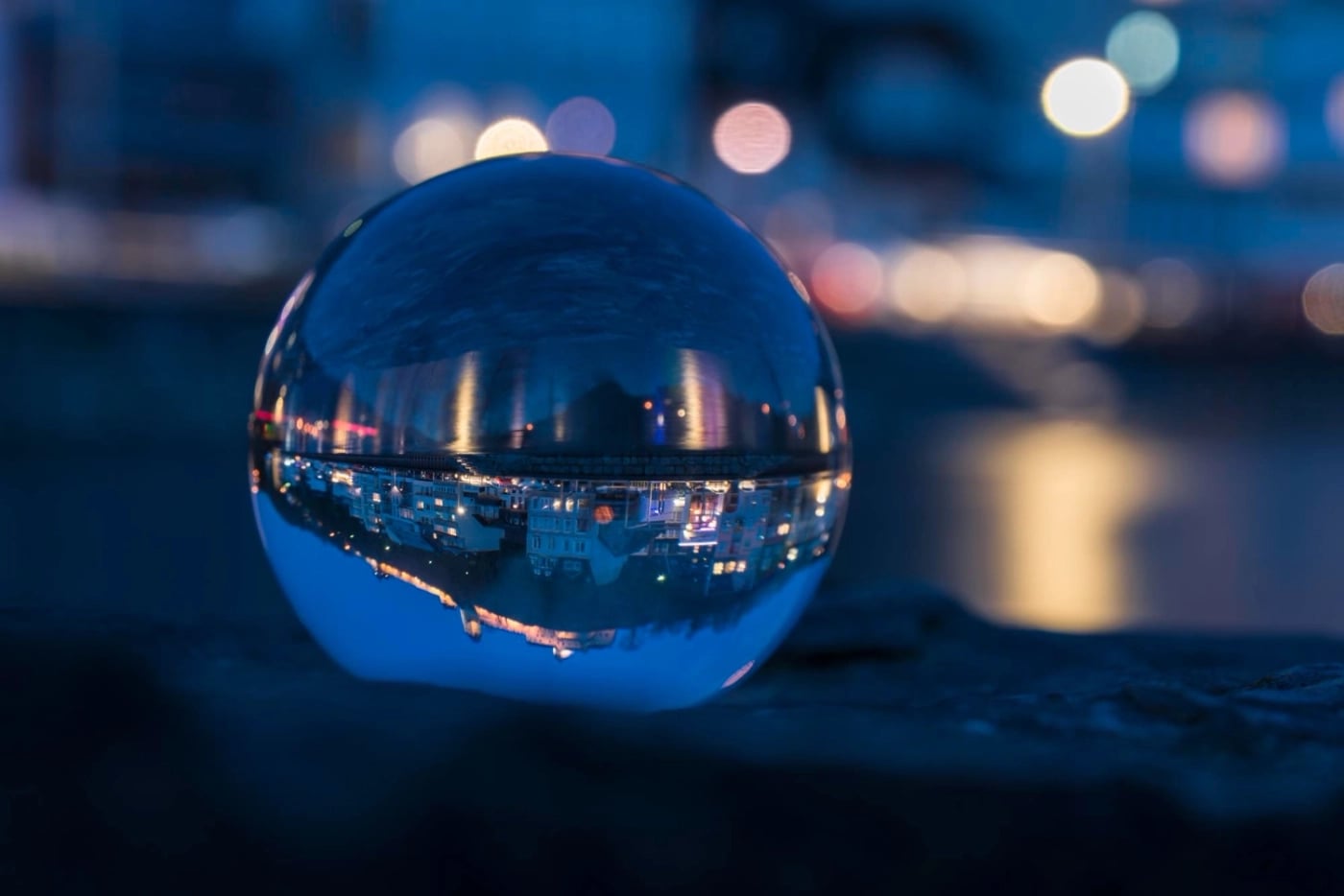 A close-up of a reflective glass sphere resting on a dark surface at night, showing an upside-down reflection of illuminated city buildings and lights across the water.
