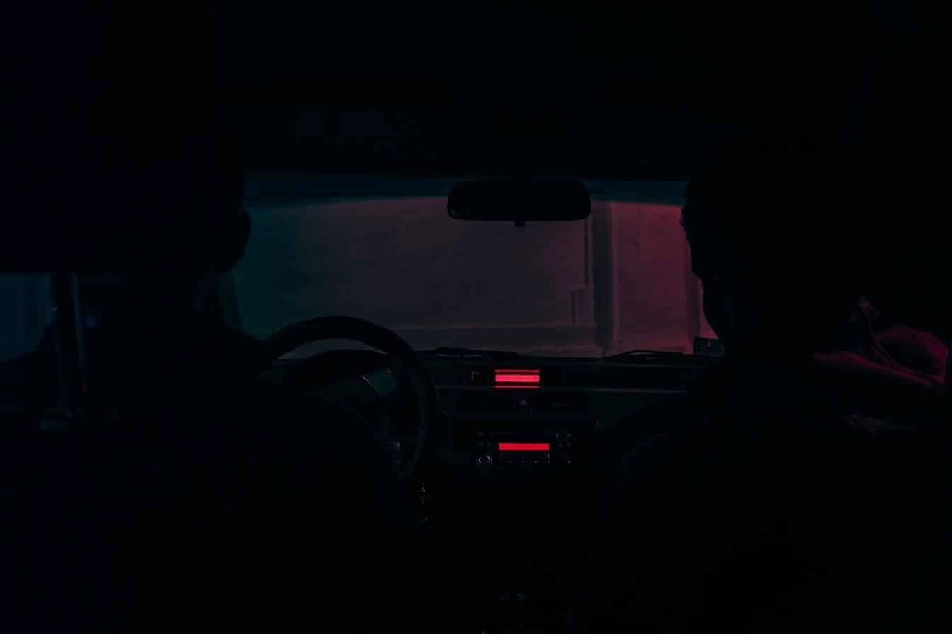 Two people sitting in a dark car interior illuminated by red and blue ambient lights through the windshield.