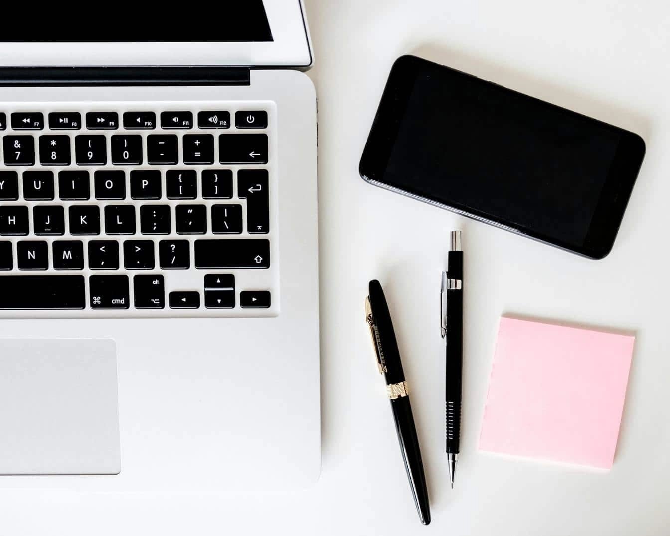 Flat lay of a laptop, smartphone, pens, and sticky notes on a white desk, representing a minimalist digital workspace for content planning or SEO tasks.