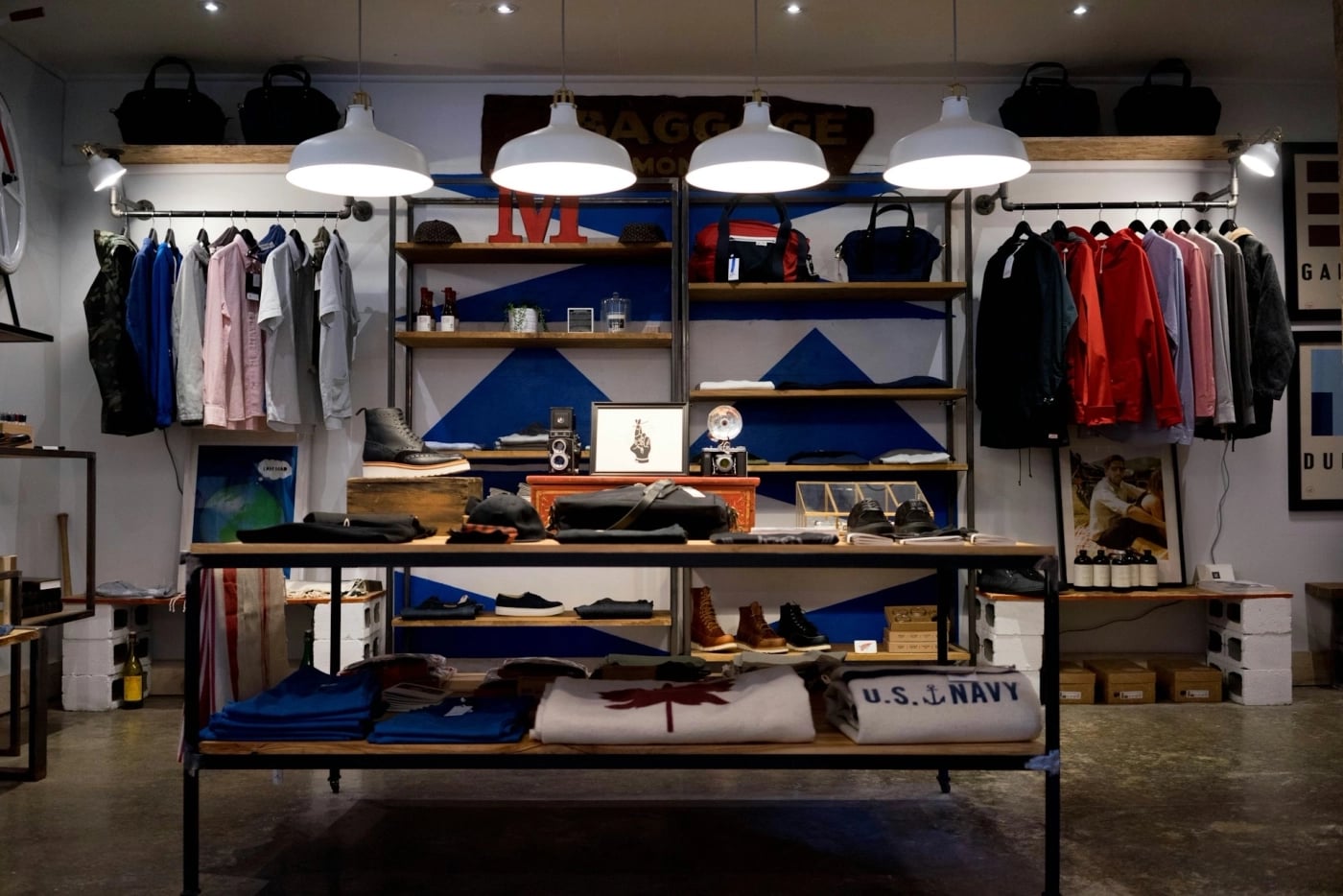 A boutique clothing store interior featuring hanging shirts, jackets, folded sweaters, shoes, and accessories neatly displayed on shelves and a central table under bright ceiling lights.