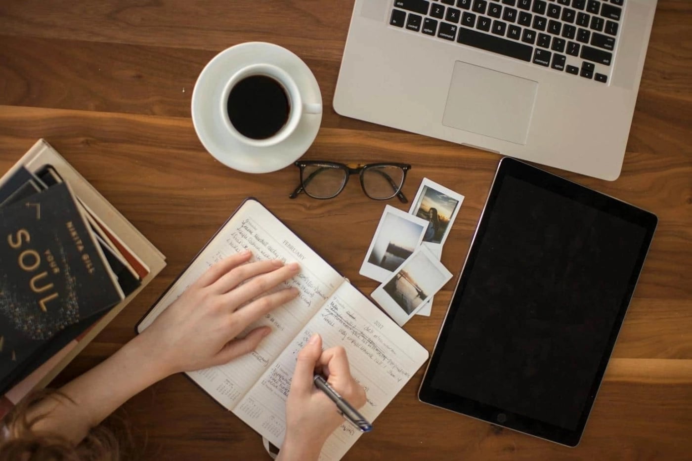 Overhead view of a workspace with a person writing in a notebook beside a laptop, tablet, coffee cup, glasses, and printed photos.