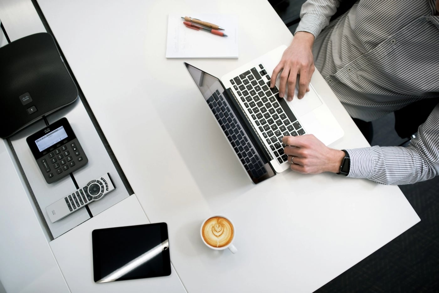Top-down view of a person in a striped shirt using a laptop at a white office desk, with a coffee cup, notepad, pens, tablet, phone, and conference speaker nearby.