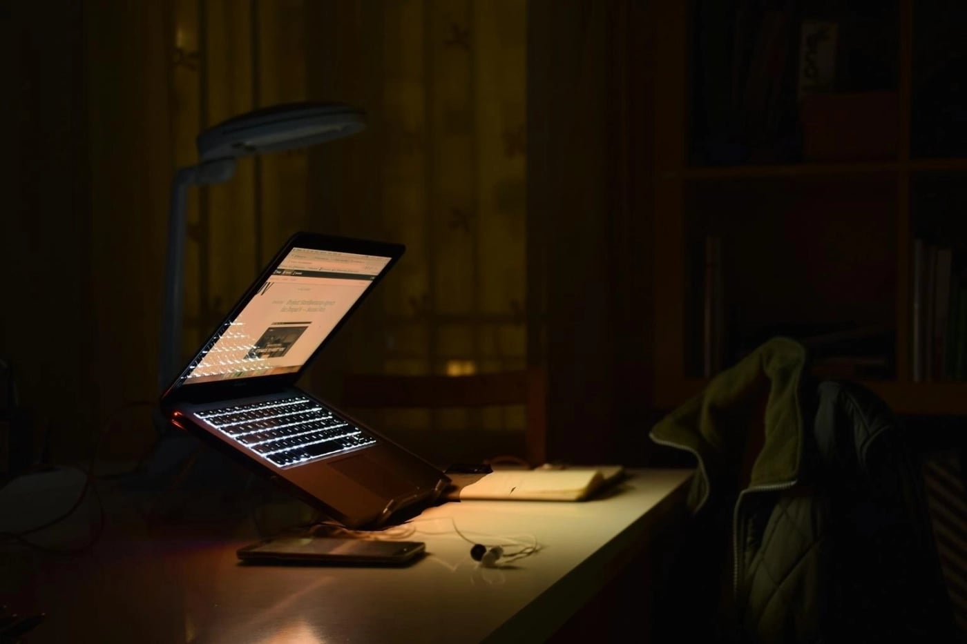 Open laptop with glowing keyboard on a desk at night, surrounded by a notebook, phone, and jacket on a chair.