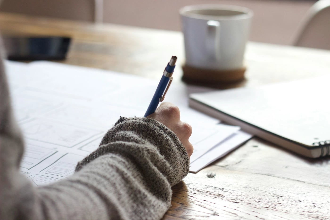 A person wearing a grey sweater writing on paper at a wooden desk with a notebook and coffee cup nearby.