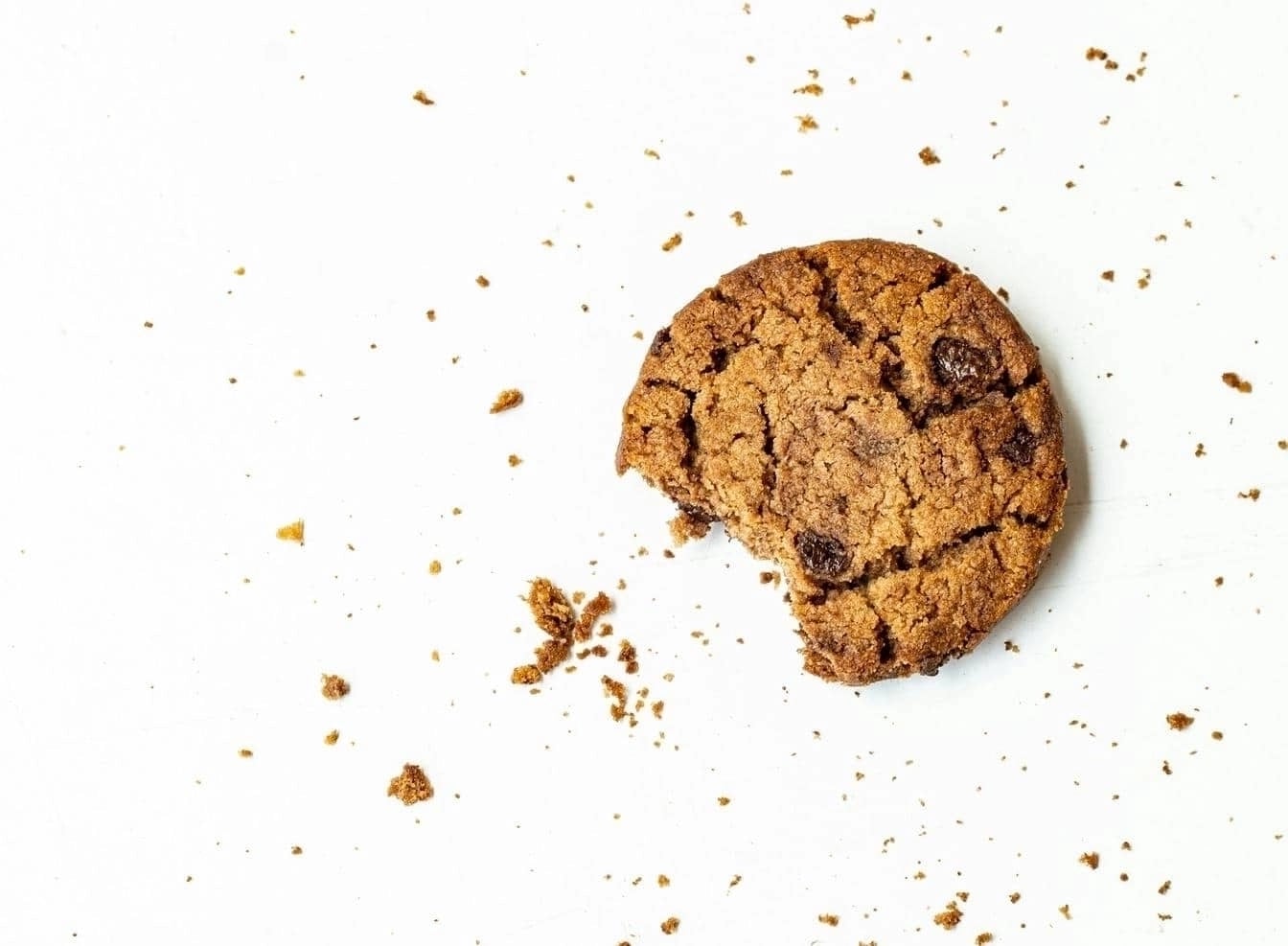 A single chocolate chip cookie with a bite taken out, resting on a white surface with scattered crumbs around it.