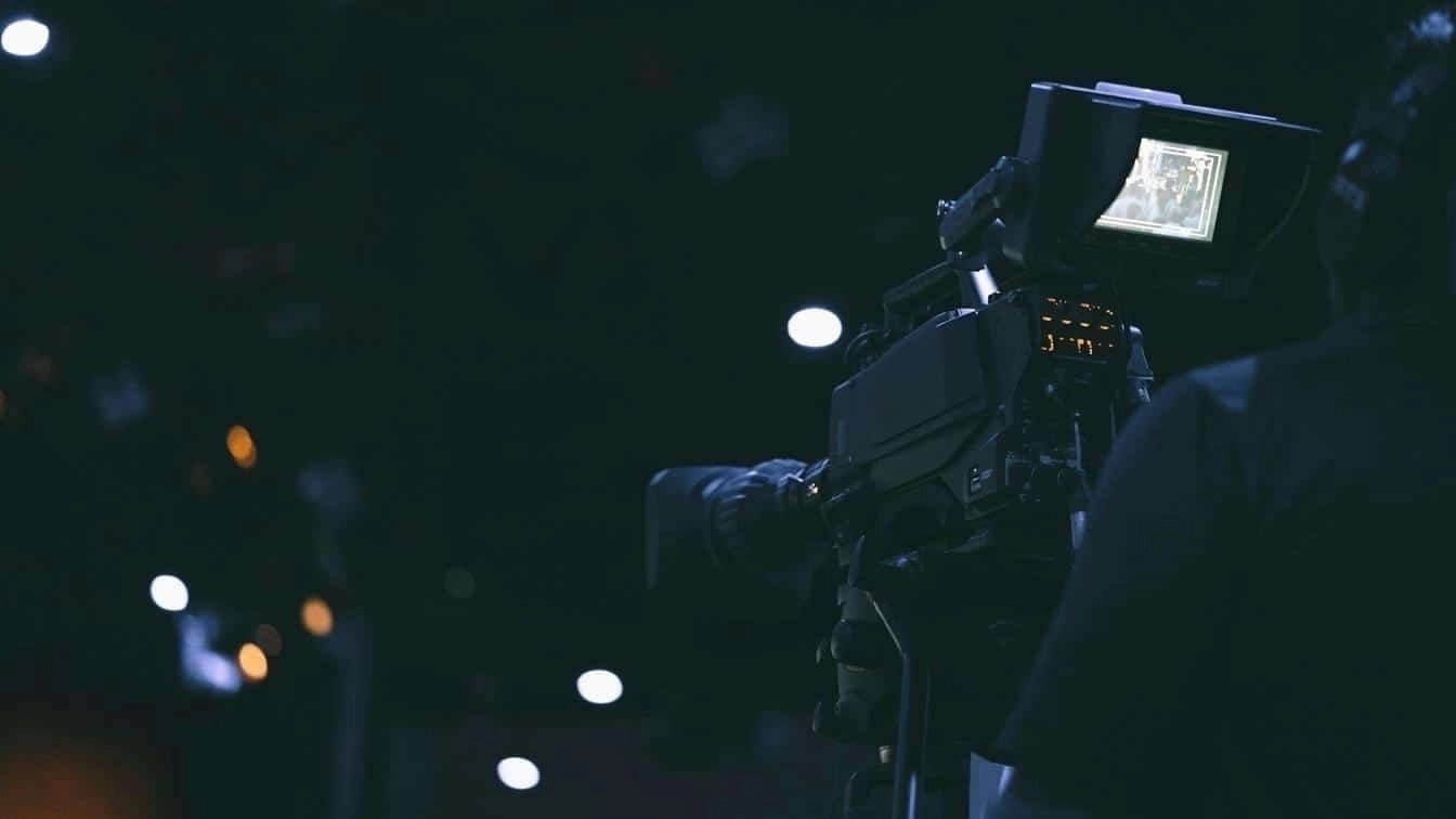 Operator behind a professional broadcast camera capturing media content in a dark studio setting.
