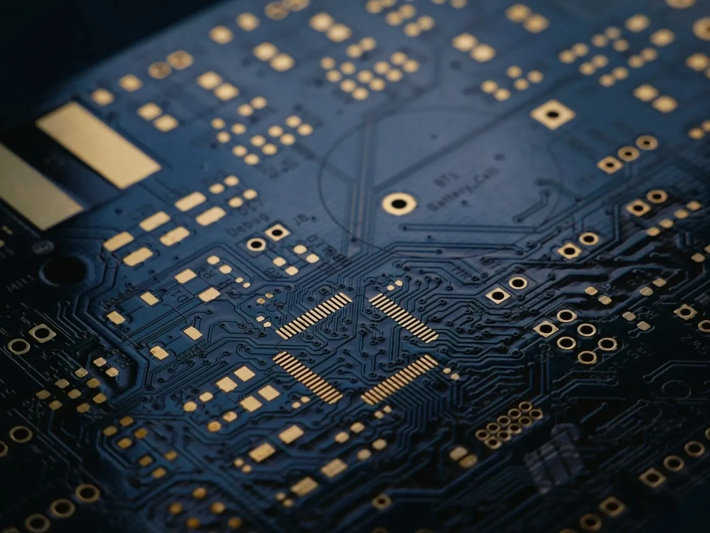 A close-up view of a printed circuit board showing intricate copper traces, solder pads, and component mounting points in gold and dark blue tones.