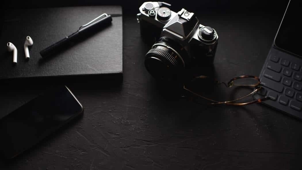 Flat lay of a camera, notebook, pen, phone, AirPods, glasses, and keyboard on a dark surface.