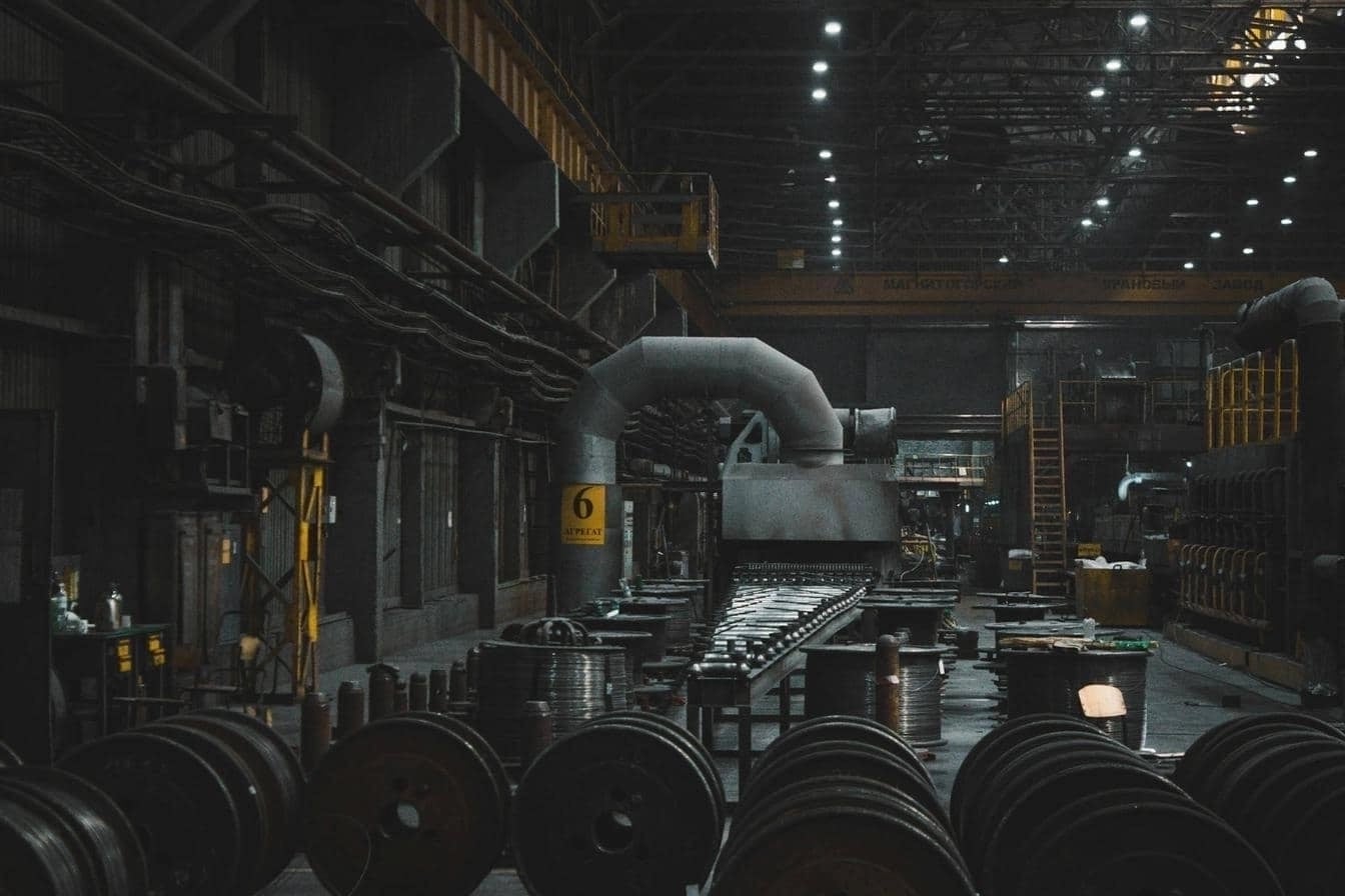 Large industrial warehouse with machinery, metal coils, ventilation systems, and production equipment under overhead lighting.
