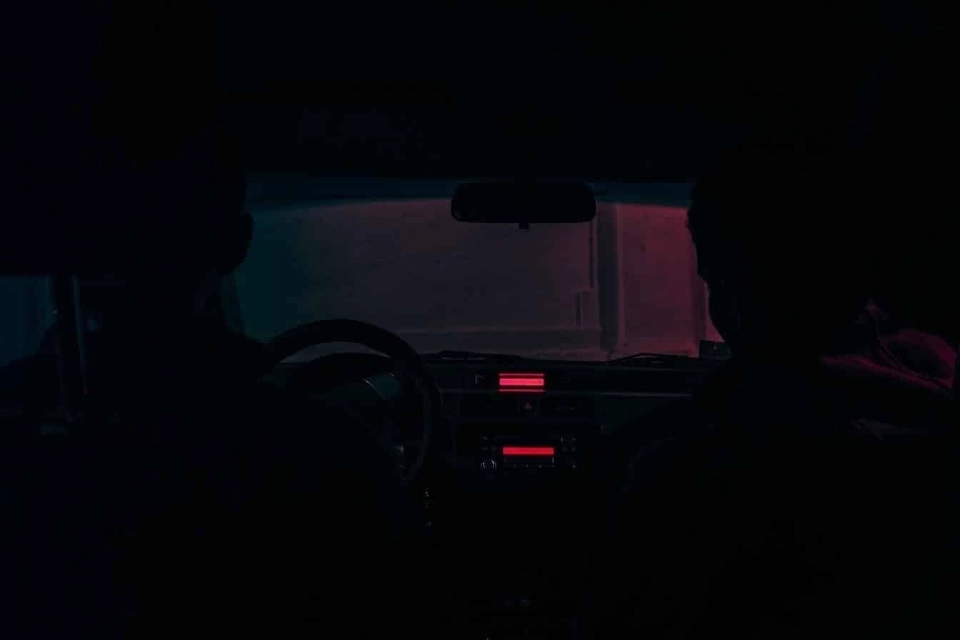 Two people sitting in a dark car interior illuminated by red and blue ambient lights through the windshield.