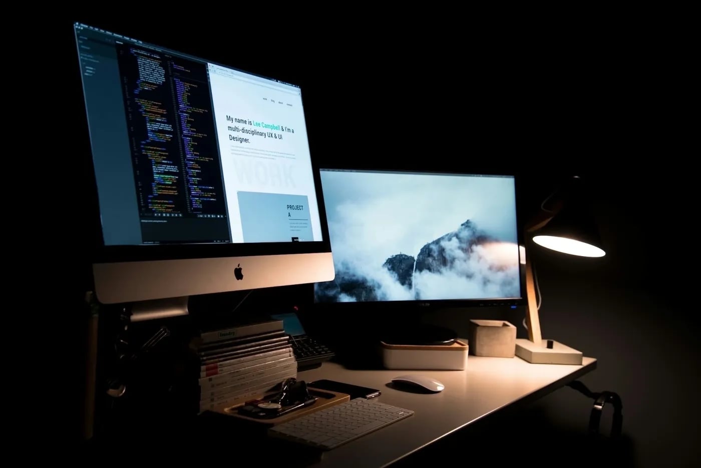 A stylish dark-themed workspace with a dual-monitor setup, displaying website code and a UX/UI design portfolio on one screen and a misty mountain wallpaper on the other, illuminated by a desk lamp.