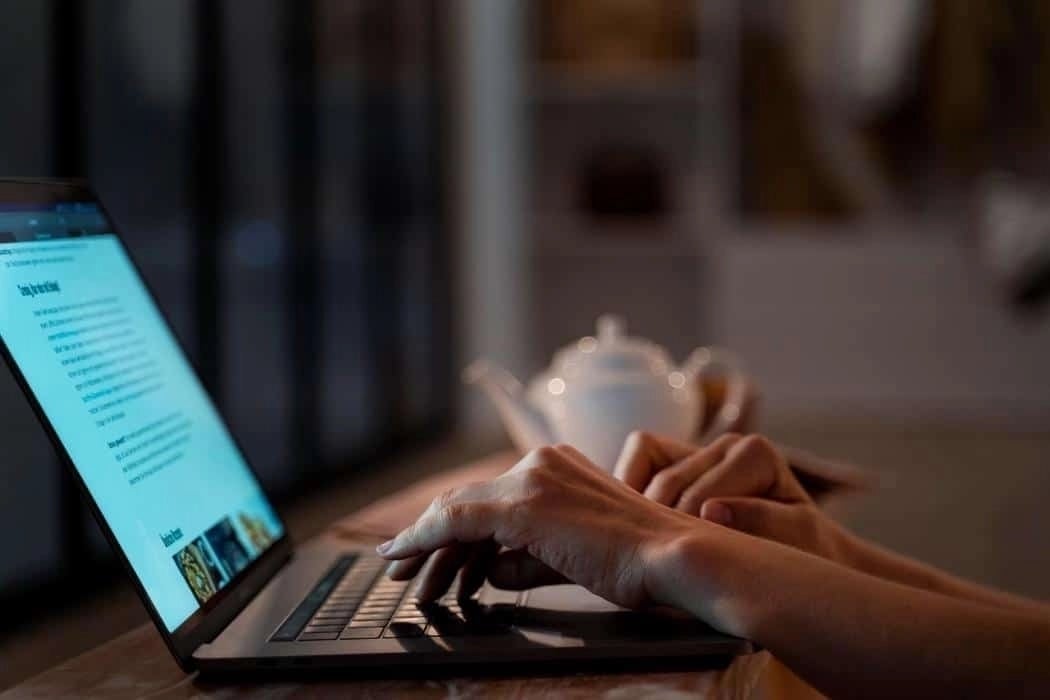 Close-up of hands typing on a laptop in a dimly lit room with a white teapot and cup in the background.