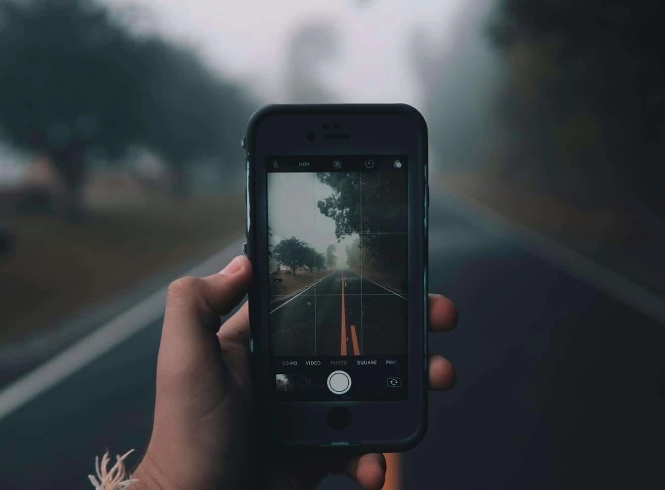 Close-up of a person holding a smartphone, capturing a photo of a foggy road lined with trees using the phone’s camera.