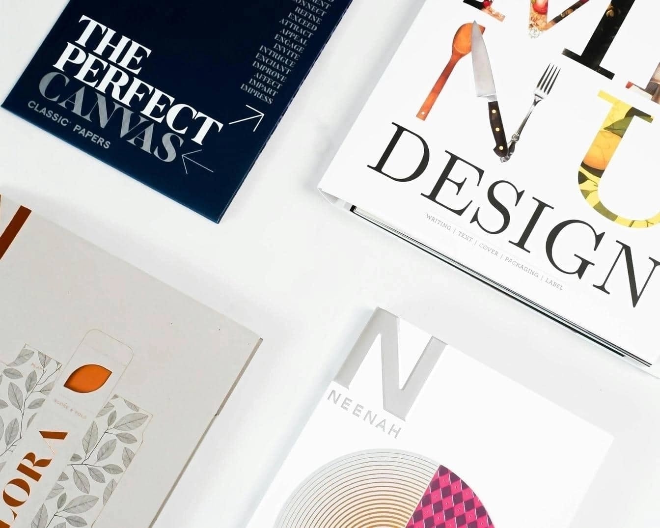Flat lay of four creative print design sample books including titles like “The Perfect Canvas,” “Design,” “Flora,” and “Neenah,” showcasing different typography and graphic styles on a white surface.