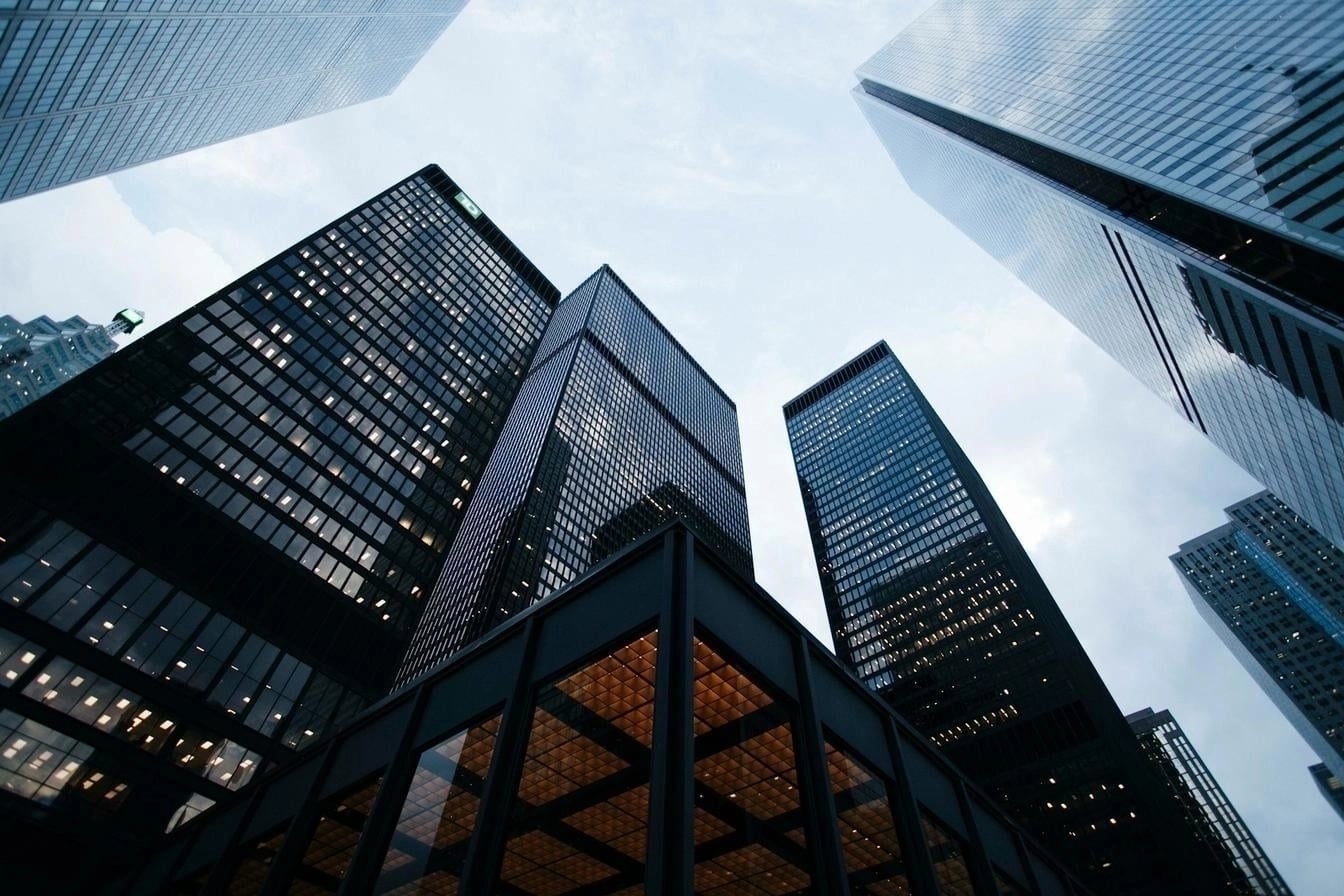 A low-angle view of tall glass skyscrapers, showcasing a modern city skyline with illuminated windows against a cloudy sky.