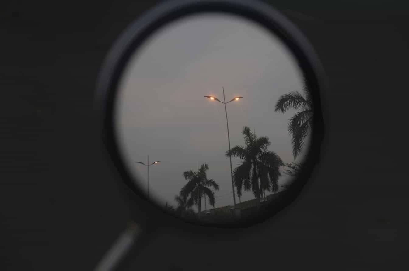 Reflection of streetlights and palm trees in a round rearview mirror against a cloudy sky.