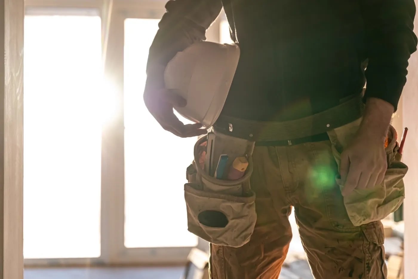 Close-up of a tradesperson holding a white hard hat with tool belt and work pants in sunlight.
