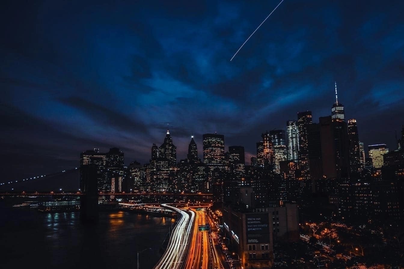 A nighttime cityscape featuring illuminated skyscrapers and long-exposure light trails from vehicles on a highway.
