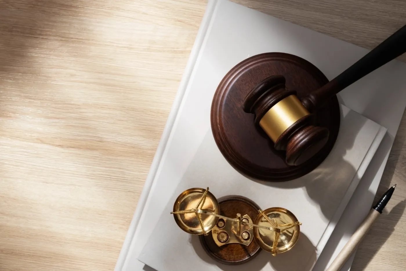 A wooden judge’s gavel with a gold band rests beside a small brass balance scale on a light wood desk, symbolising law and justice.