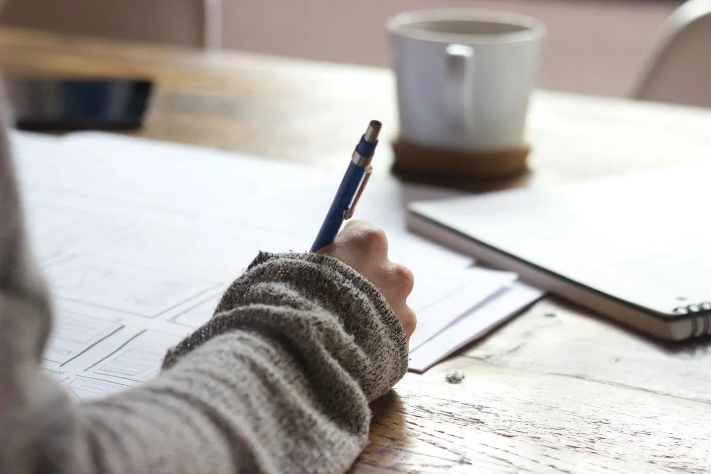 A person wearing a grey sweater writing on paper at a wooden desk with a notebook and coffee cup nearby.