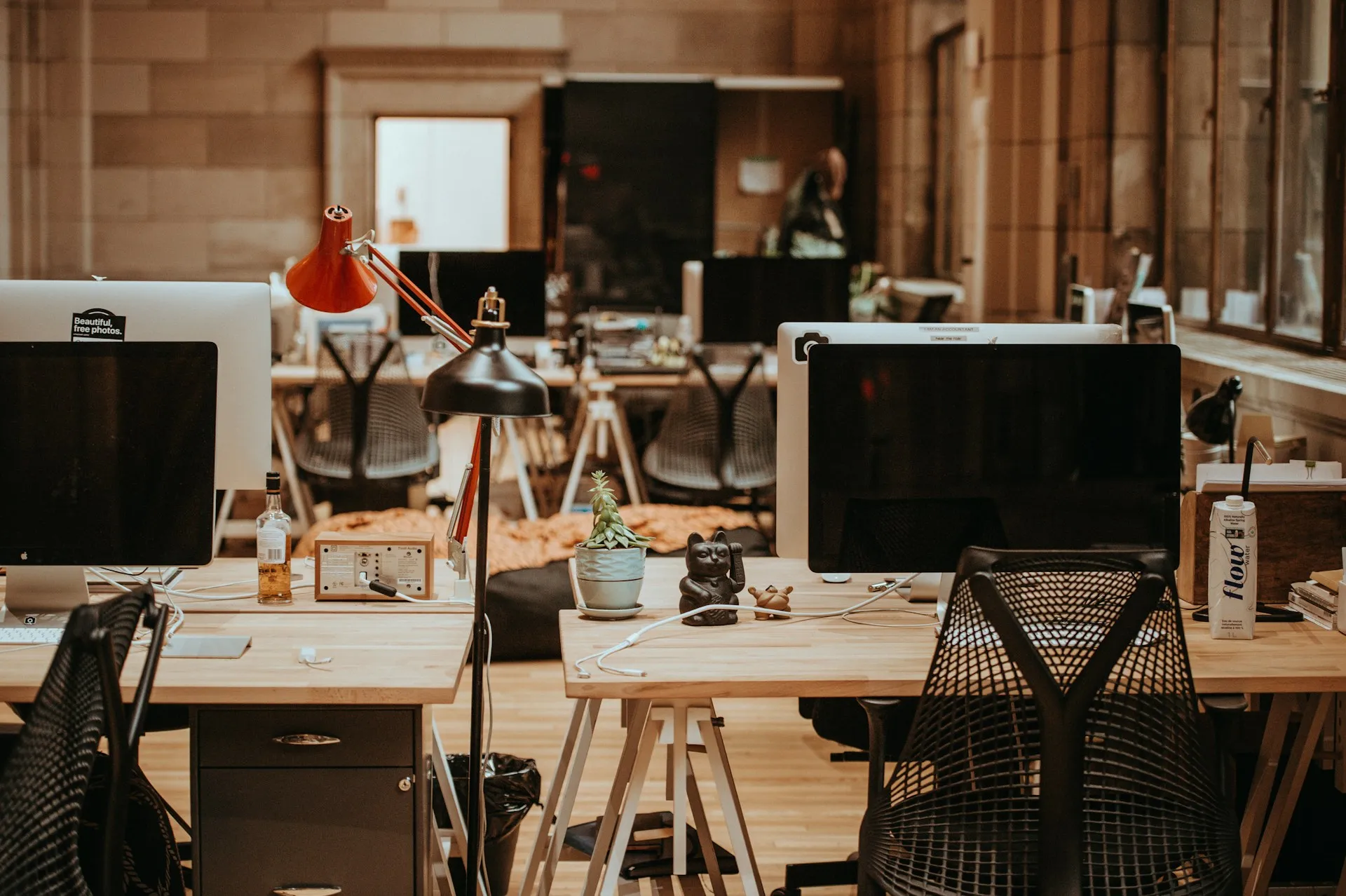 Modern open-plan office with wooden desks, iMacs, desk lamps, and various personal items like a potted plant and lucky cat figurine.