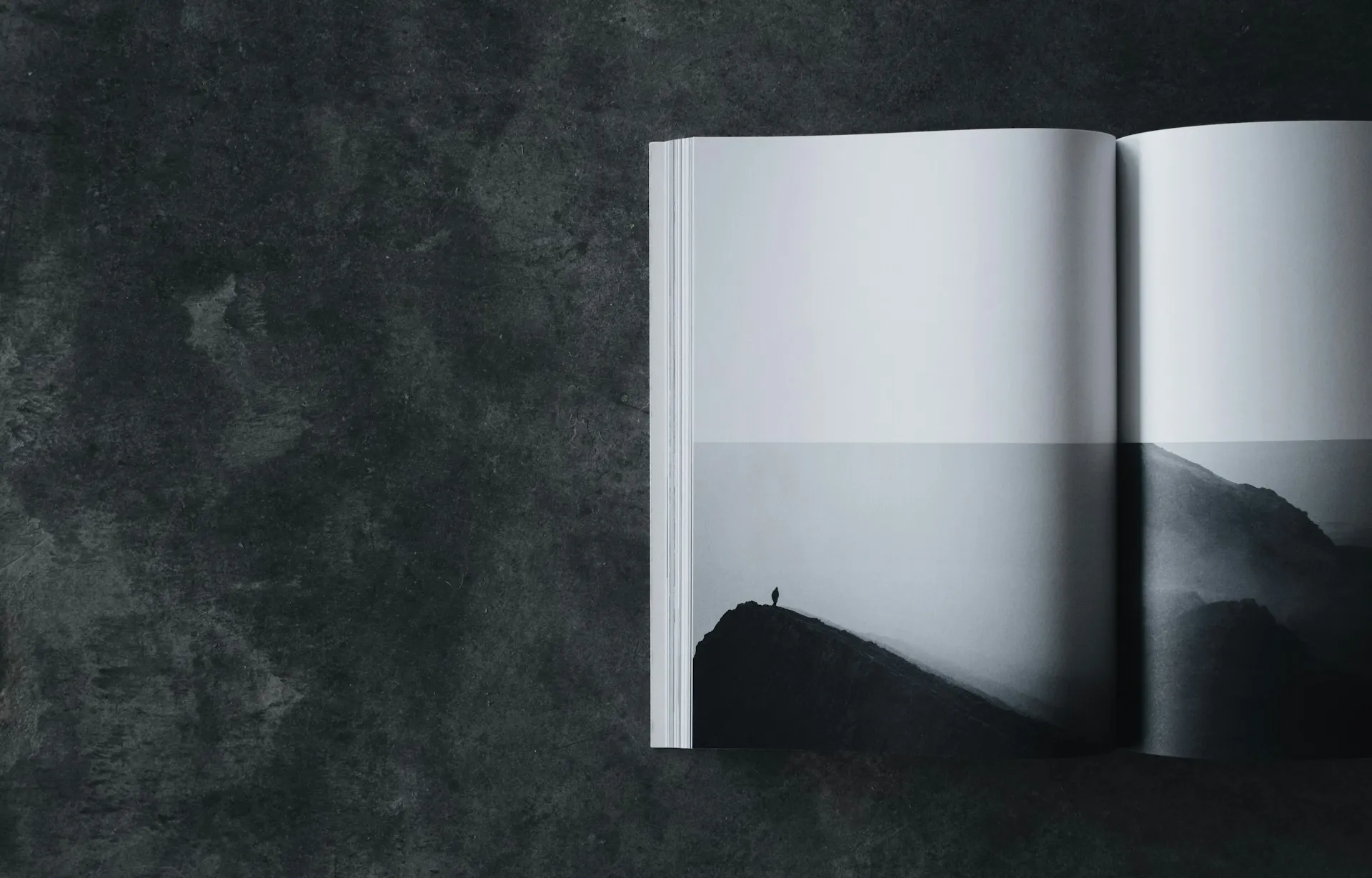 Open book displaying a black and white photo of a lone figure on a mountain ridge, placed on a dark textured surface.