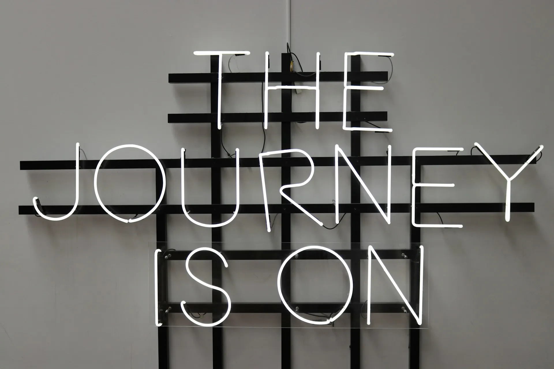 Neon sign reading 'THE JOURNEY IS ON' mounted on a minimalist black grid against a light grey wall.