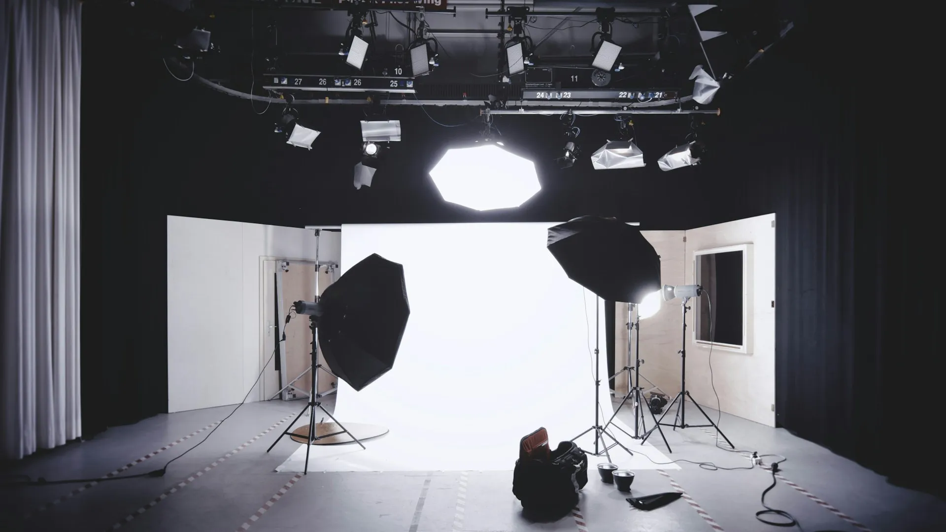 Professional photo studio setup with large softbox lights, umbrellas, and a white backdrop surrounded by overhead lighting equipment.