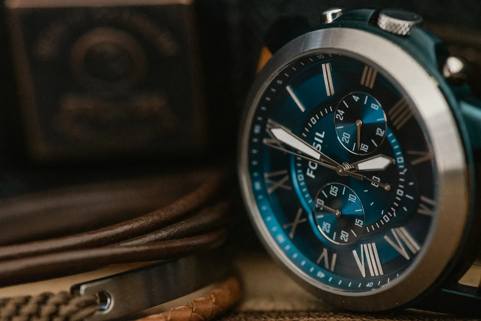 Stylish men's wristwatch with a blue face and Roman numerals, placed on a leather surface with accessories.