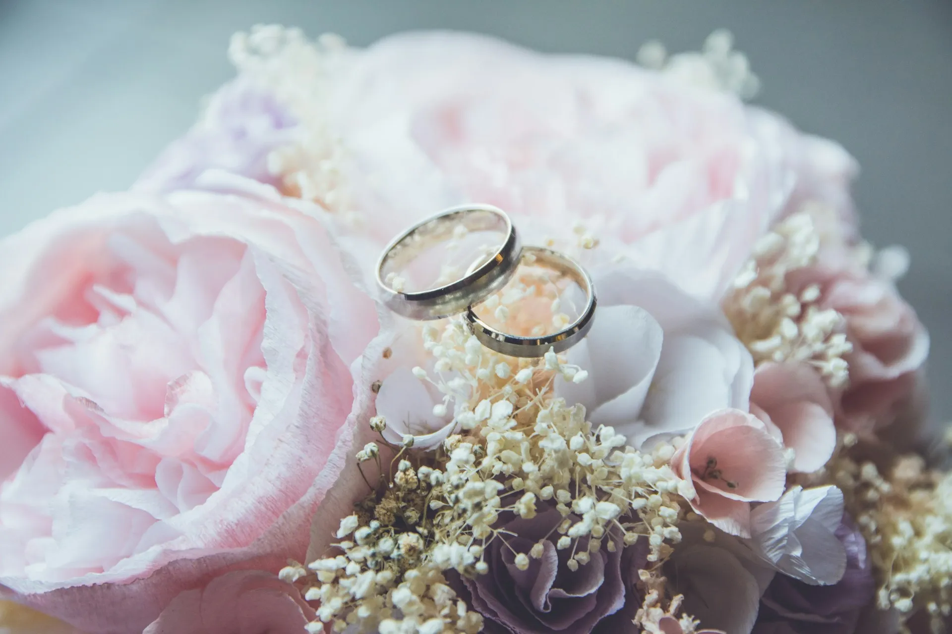 Two silver wedding rings resting on a soft pastel bouquet of roses, baby's breath, and other delicate flowers.