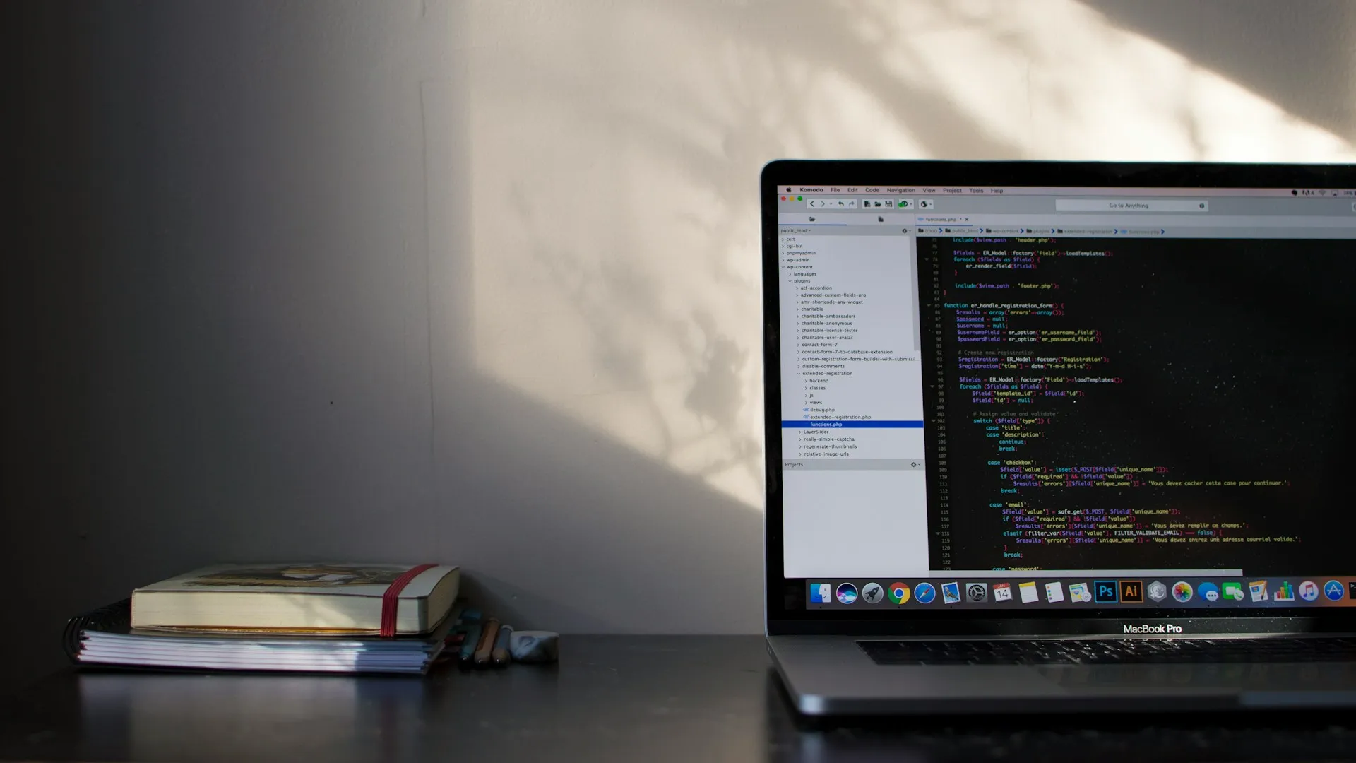 MacBook Pro on a desk displaying code in a development environment, next to a stack of notebooks and pencils with soft sunlight casting shadows on the wall.