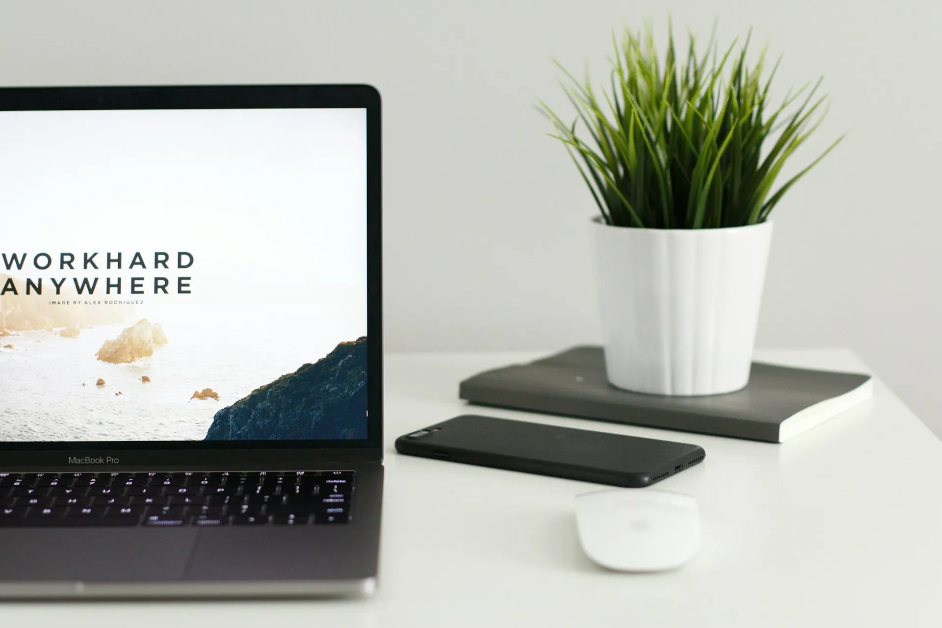 Minimalist workspace with a MacBook Pro displaying “Work Hard Anywhere,” alongside a potted plant, smartphone, mouse, and notebook on a white desk.
