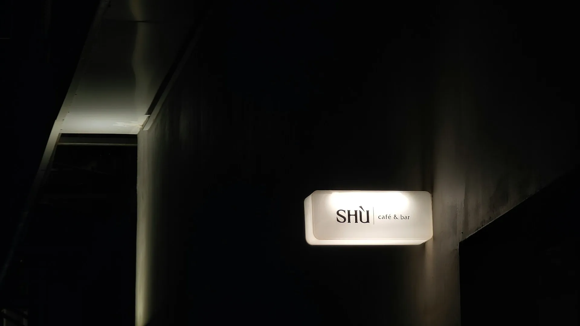 Dimly lit corridor with an illuminated sign reading 'SHÙ café & bar' mounted on a dark wall.