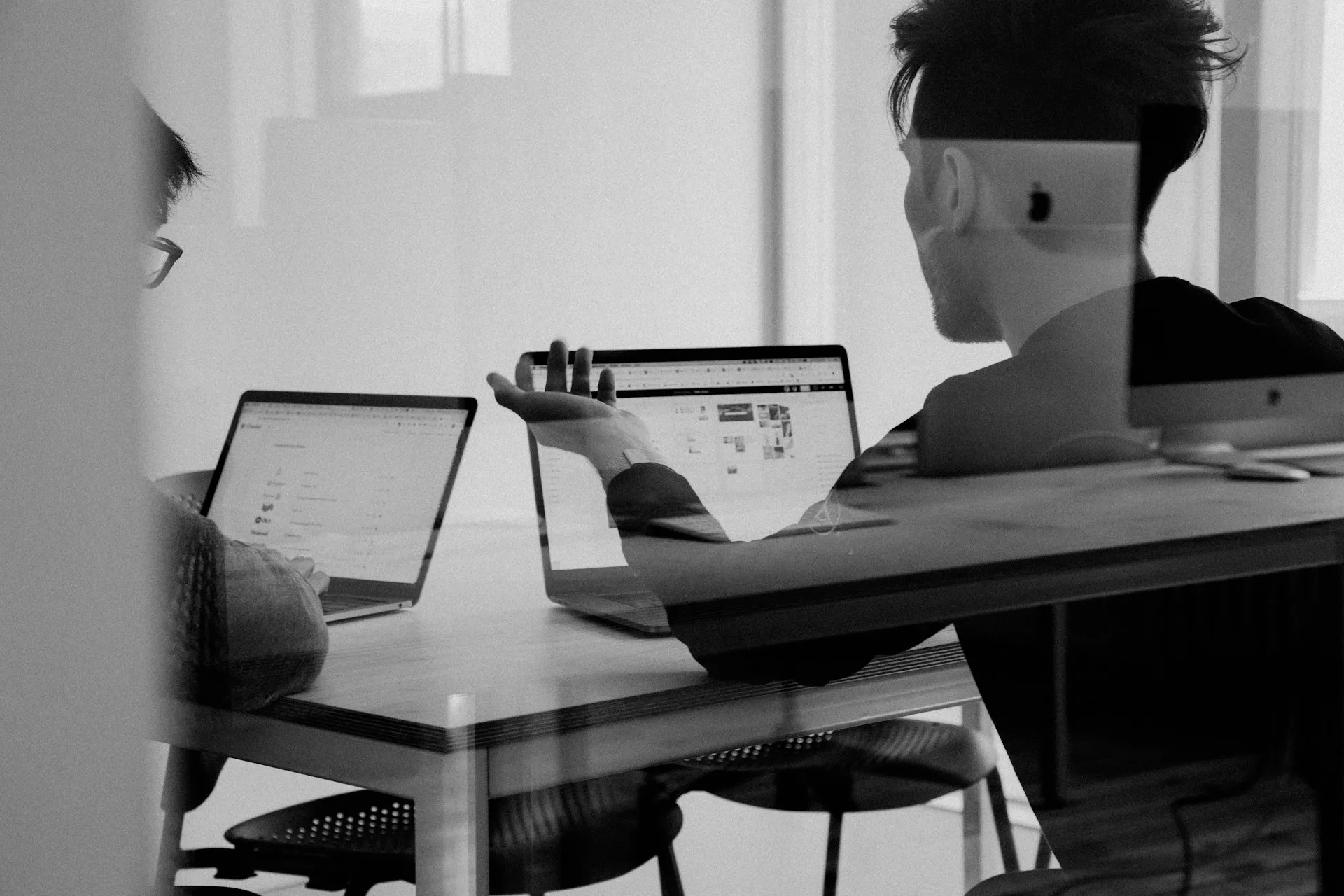 Two people collaborating at a desk Two people collaborating at a desk with laptops, viewed through a glass partition, in a modern office setting.