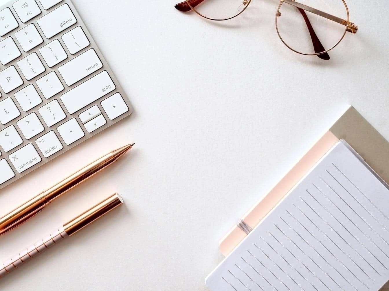 Flat lay of office desk with keyboard, glasses, gold pen, and lined notepad.