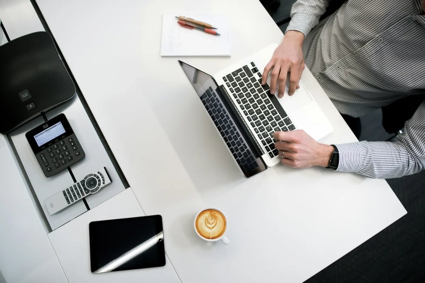 Top-down view of a person in a striped shirt using a laptop at a white office desk, with a coffee cup, notepad, pens, tablet, phone, and conference speaker nearby.